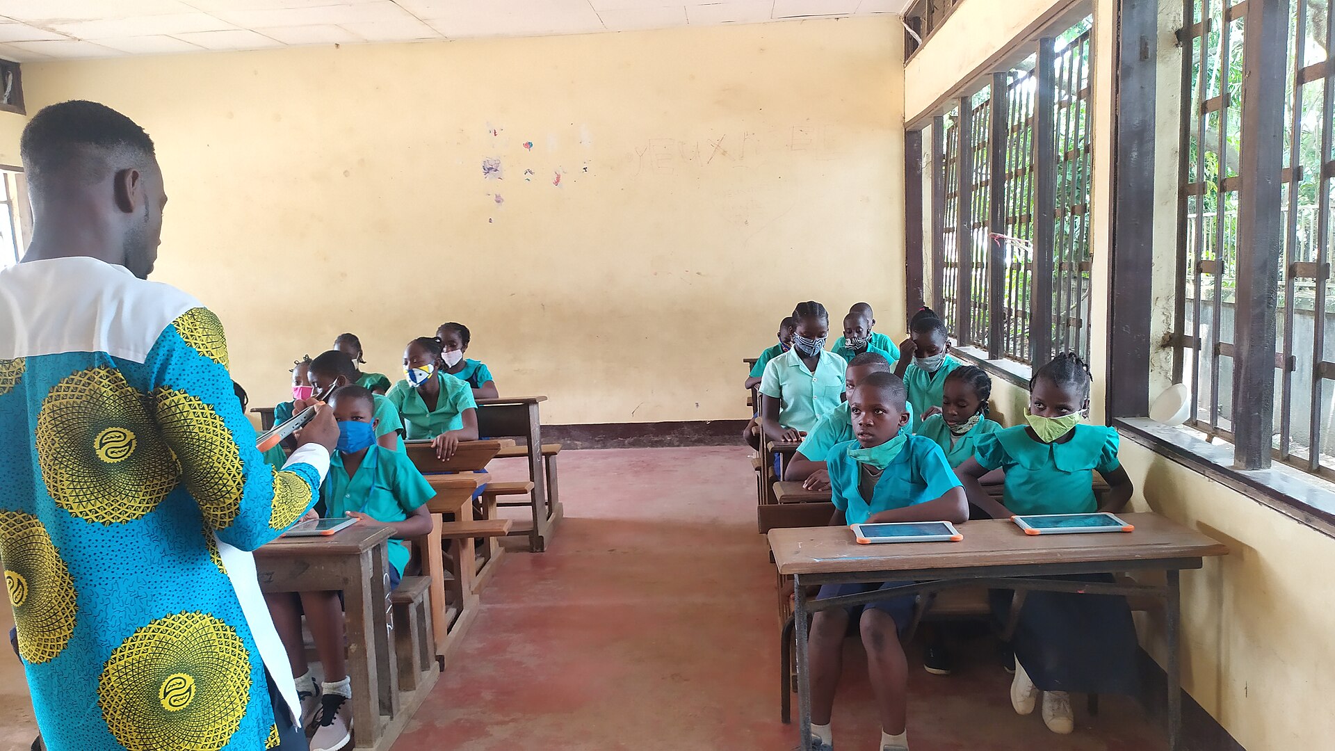 Pupils in green school uniforms at their desks while a teacher in a bright African print shirt speaks to the class