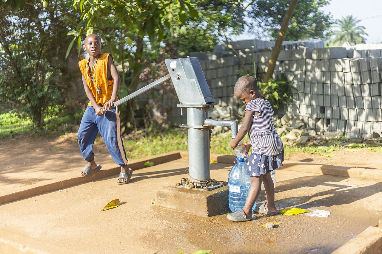Two children collecting water from a hand pump on a sunny morning